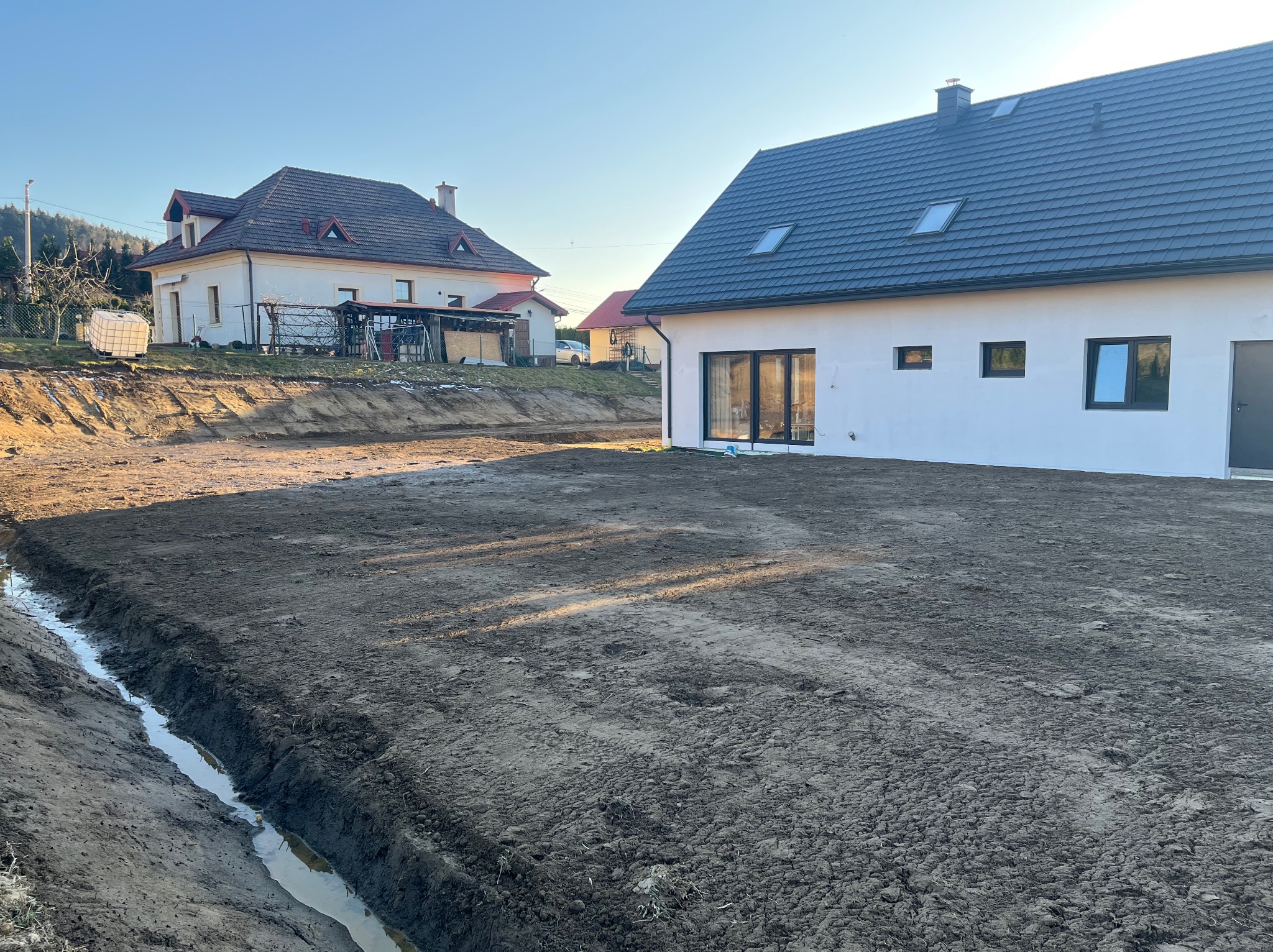 Przygotowany teren pod budowę z wykopanym rowem melioracyjnym, widoczne dwa domy w tle, wczesne popołudnie, słoneczna pogoda.
