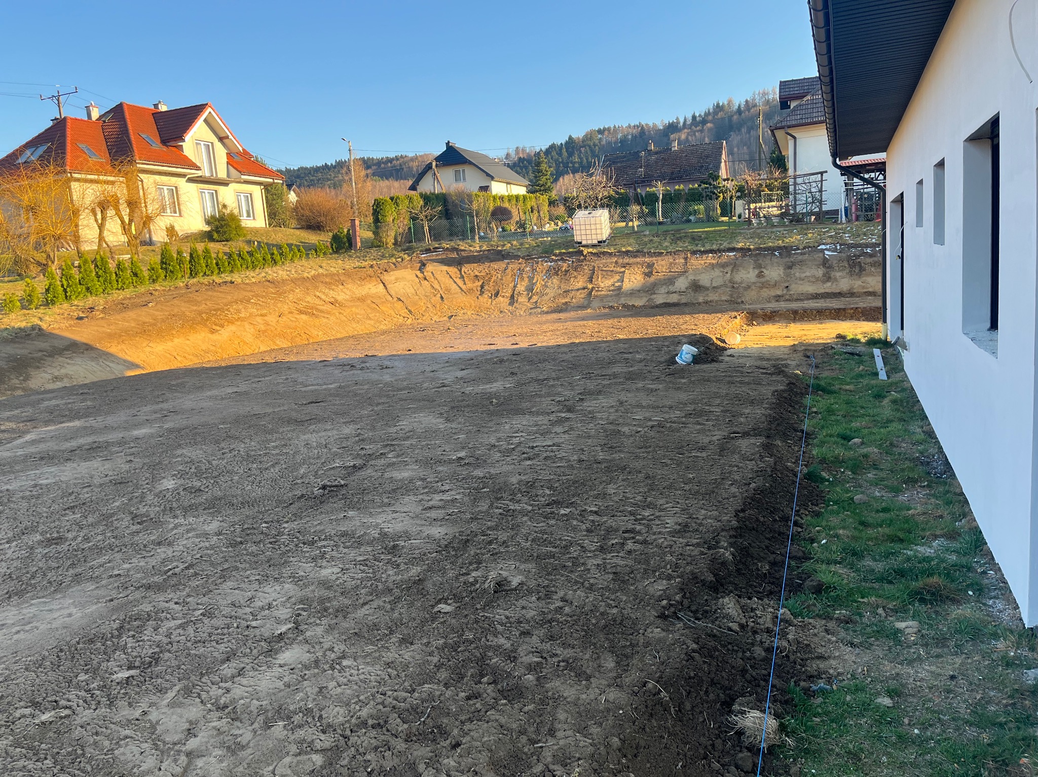 Wykop ziemny pod budowę, widok z boku budynku z oknami. W tle domy i drzewa w słoneczny dzień. Wykop zabezpieczony sznurkiem.