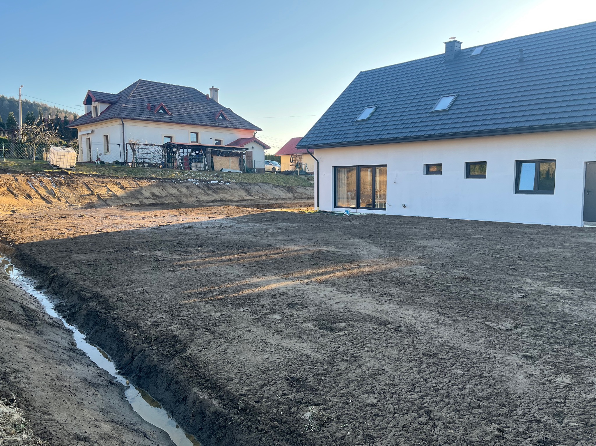 Wykonany drenaż liniowy z wodą, obok nowo wybudowany dom z czarnymi oknami i szarą dachówką, w tle drugi dom. Widok na posesję po wykonanych pracach ziemnych.