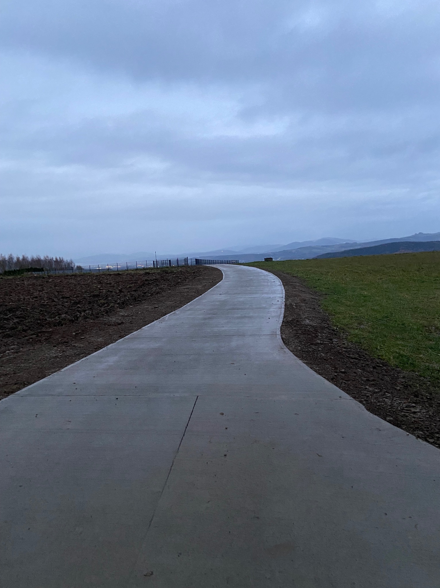 Betonowa ścieżka wijąca się przez krajobraz Bartkowa-Posadowa, z widokiem na wzgórza i pochmurne niebo. Ścieżka łączy pole uprawne z trawiastym terenem.