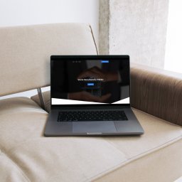 PROJSTOG Oscar Grzywa - Strony Internetowe - Laptop na sofie prezentuje stronę www 'Strefa Nieruchomości Mielec'. Minimalistyczny design, stonowana kolorystyka. Widok z bliska, naturalne światło.