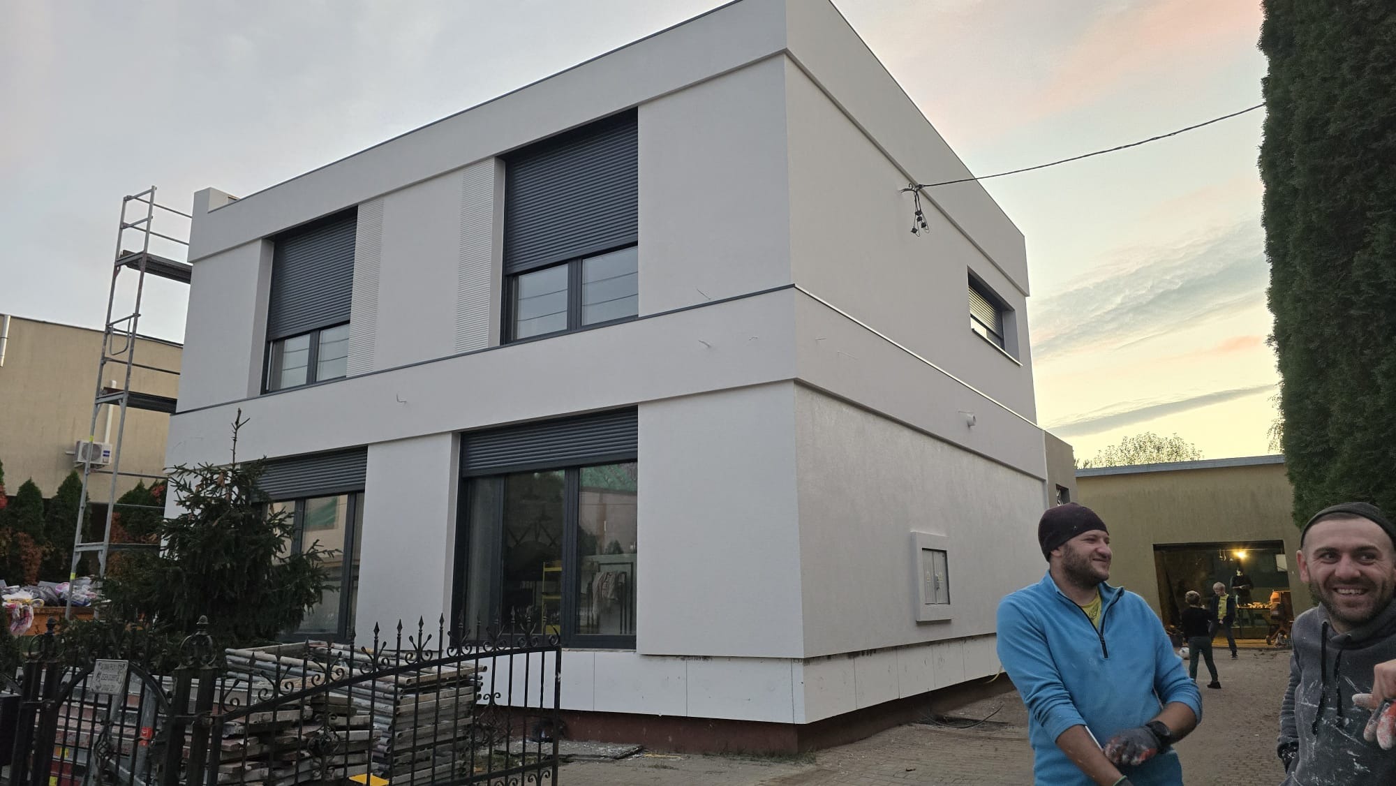 Nowoczesny dom z szarymi roletami zewnętrznymi, świeżo po remoncie elewacji. Na pierwszym planie pracownicy w trakcie sprzątania po pracy.