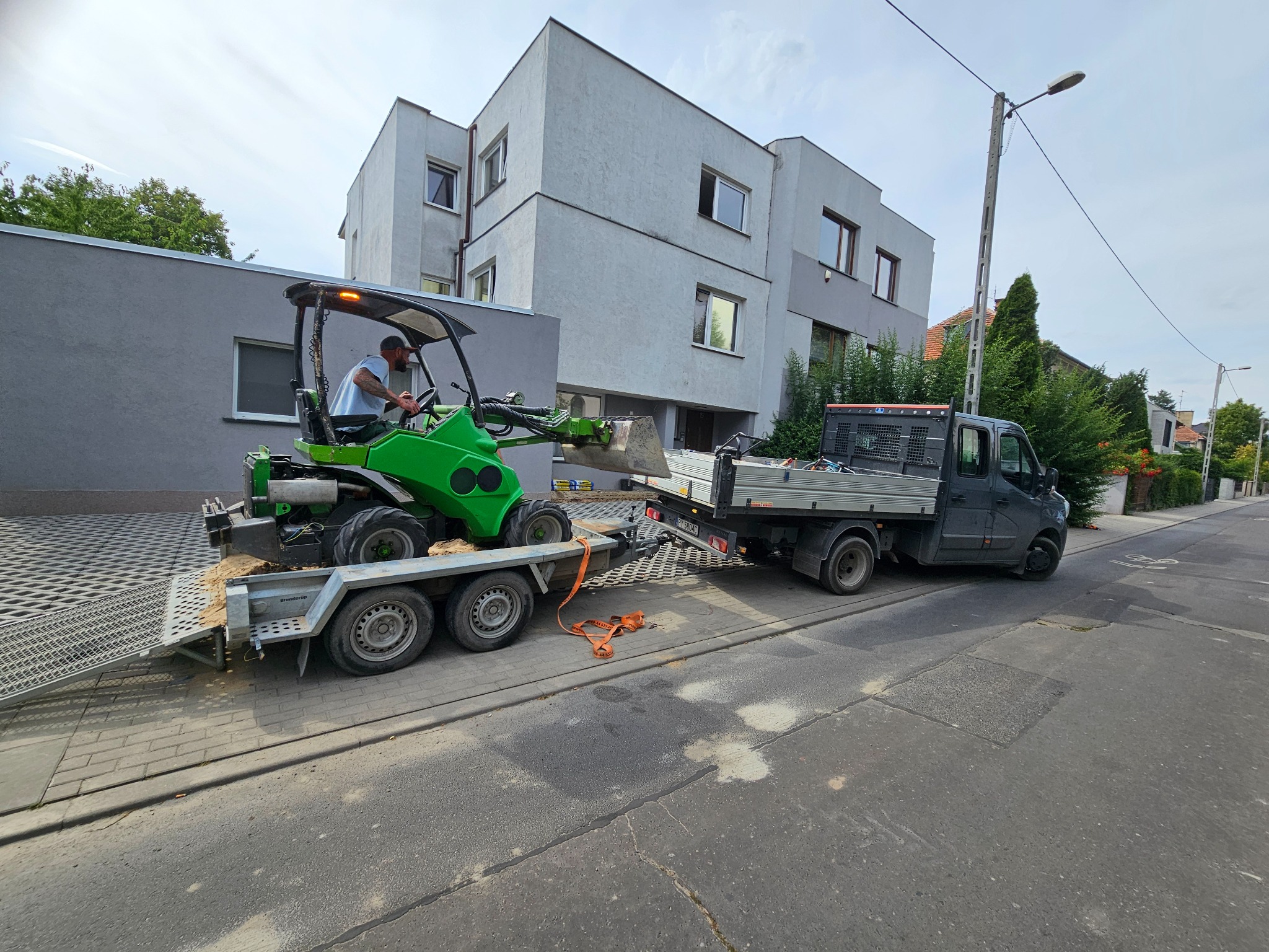 Zielona mini koparka na przyczepie, ciągnięta przez szary samochód dostawczy na tle jasnego budynku. Operator w kabinie. Kostka brukowa na podłożu.