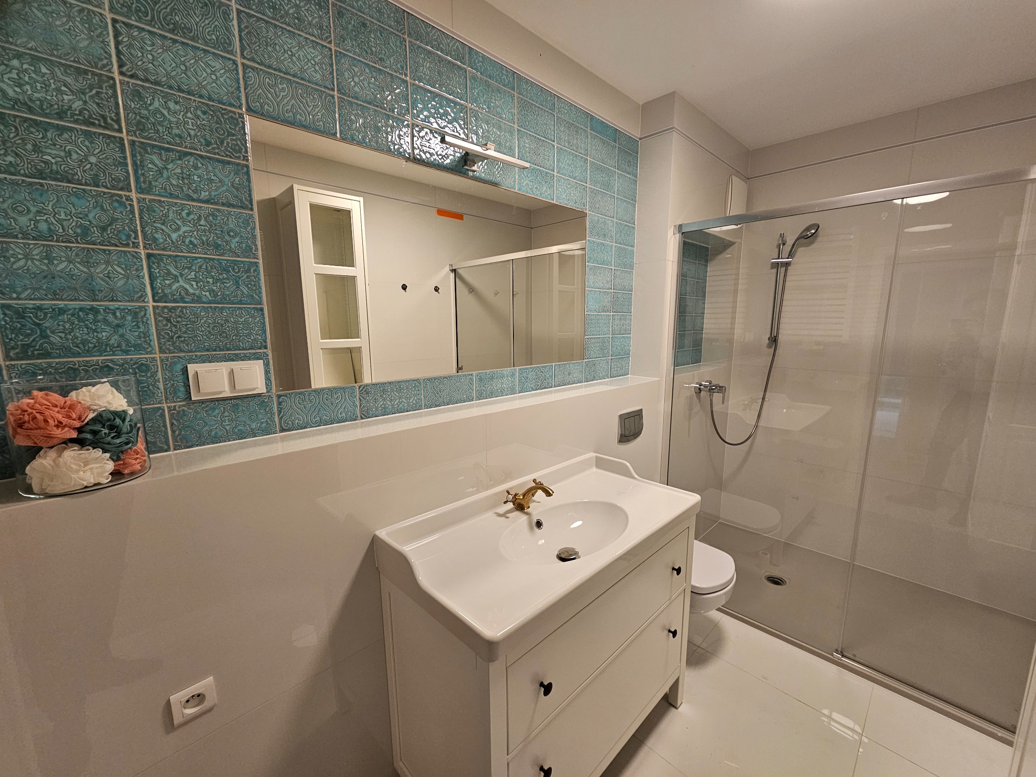 Biała szafka pod umywalkę z ozdobną baterią, turkusowe płytki na ścianie i kabina prysznicowa. Widok na odnowioną łazienkę z lustrem i akcesoriami.
