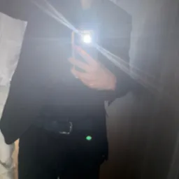 Mężczyzna w czarnym garniturze robi selfie w lustrze, widoczny fragment twarzy, ręka z telefonem i odbicie sylwetki. Silne odbicie światła z telefonu.