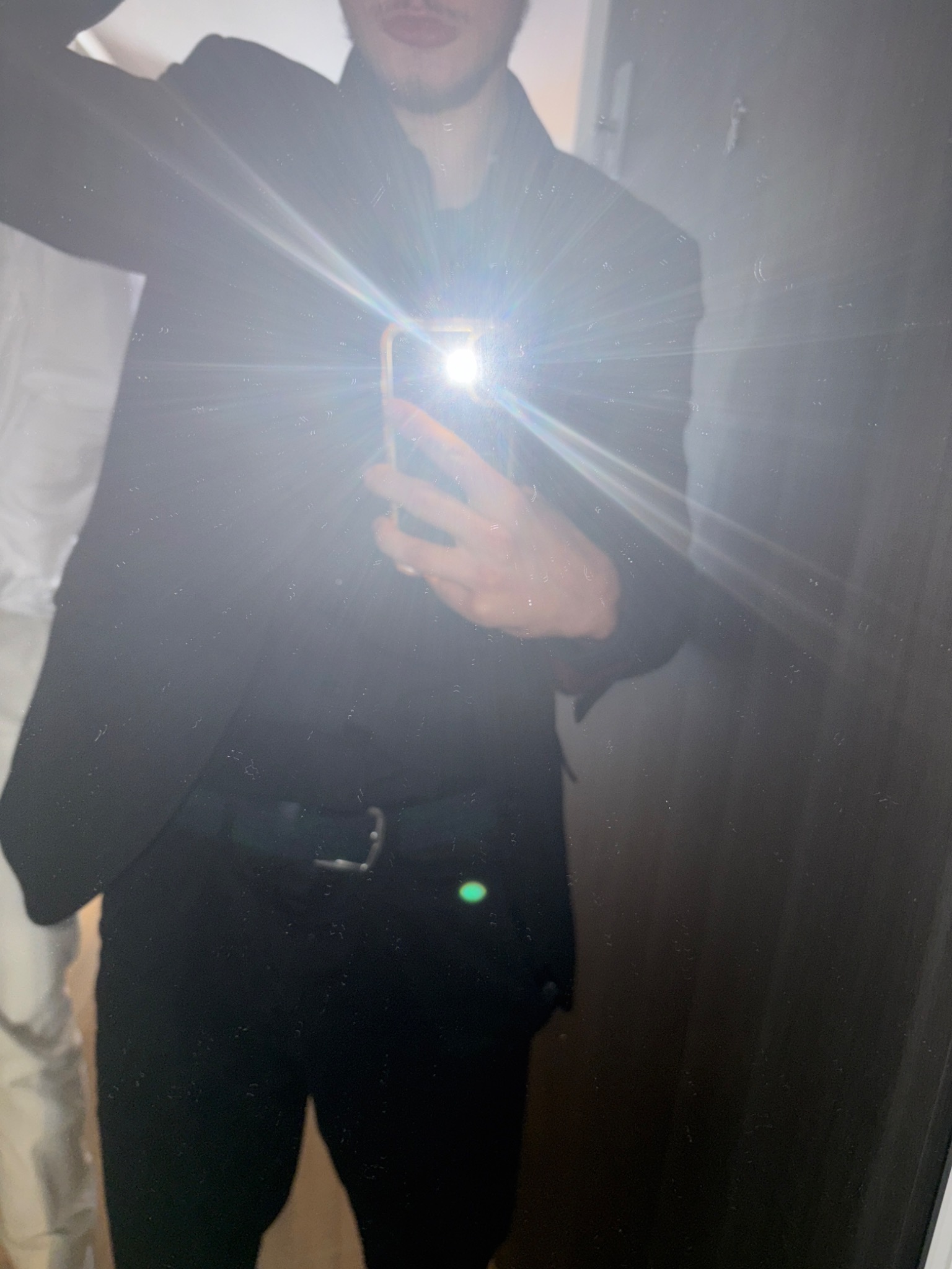 Mężczyzna w czarnym garniturze robi selfie w lustrze, widoczny fragment twarzy, ręka z telefonem i odbicie sylwetki. Silne odbicie światła z telefonu.