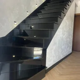 Eleganckie, czarne schody kamienne z subtelnym oświetleniem punktowym wzdłuż ściany, harmonizujące z parkietem ułożonym w jodełkę.