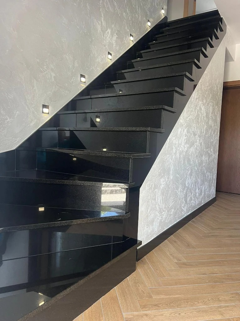 Eleganckie, czarne schody kamienne z subtelnym oświetleniem punktowym wzdłuż ściany, harmonizujące z parkietem ułożonym w jodełkę.