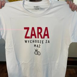 ProPunktBHP - Biała koszulka z czerwonym napisem 'ZARA Wychodzę za mąż' i grafiką obrączek, prezentowana na tle ściany z panelami akustycznymi i fragmentem podłogi.