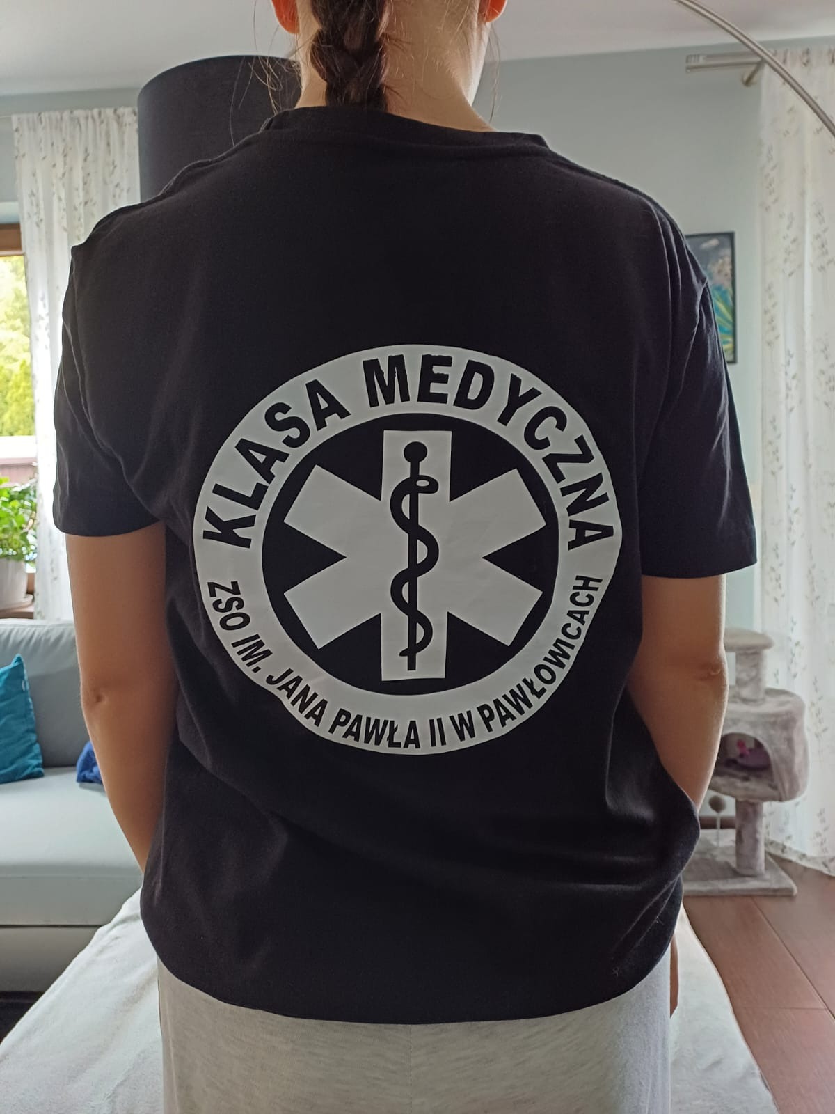 Osoba w czarnej koszulce z białym nadrukiem: gwiazda życia i napis 'KLASA MEDYCZNA ZSO IM. JANA PAWŁA II W PAWŁOWICACH'. Widok z tyłu, w tle wnętrze.