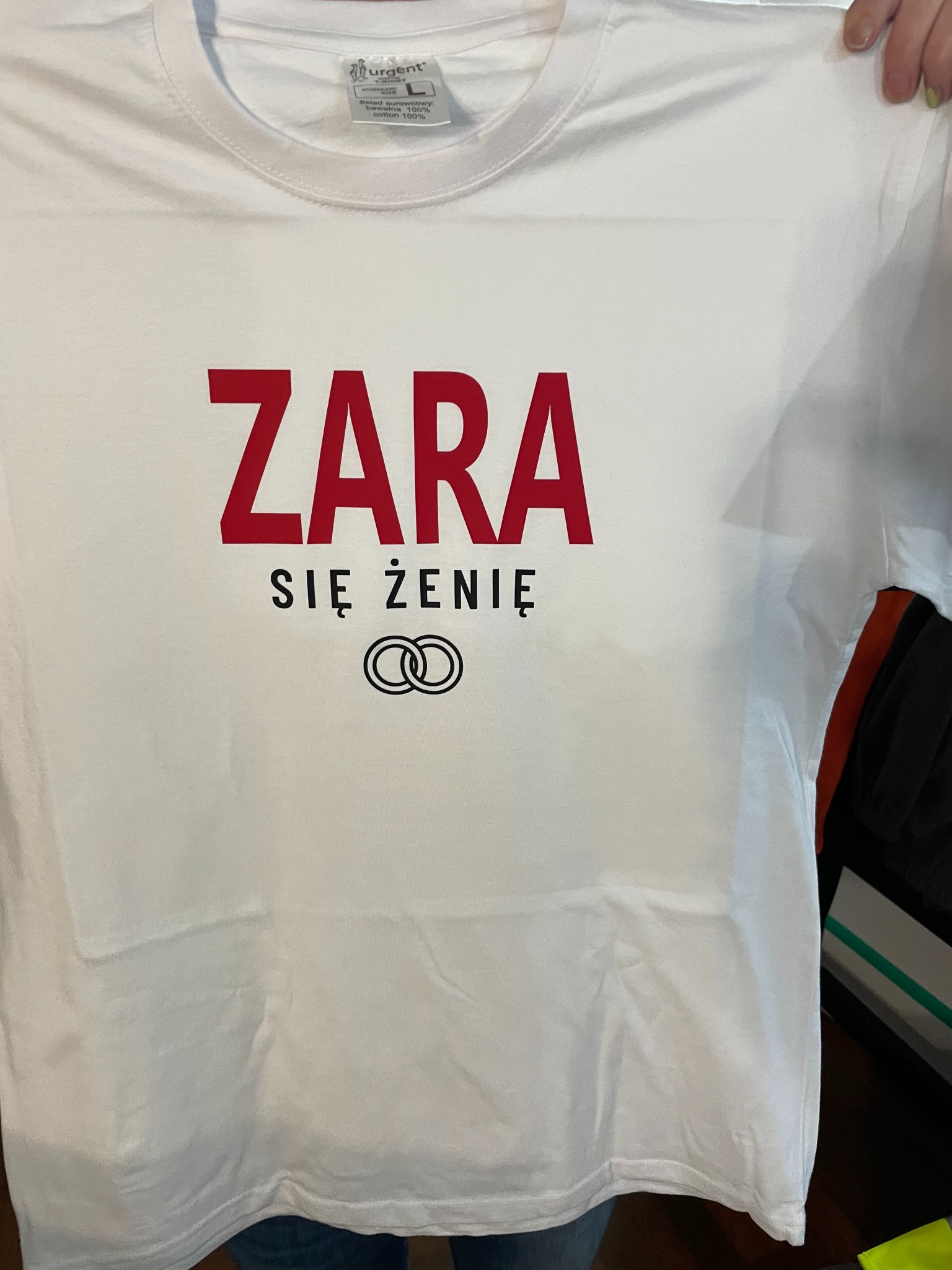 Biała koszulka z czerwonym napisem 'ZARA SIĘ ŻENIĘ' i symbolem obrączek. Prezent dla przyszłego pana młodego. Widoczna metka producenta Urgent.