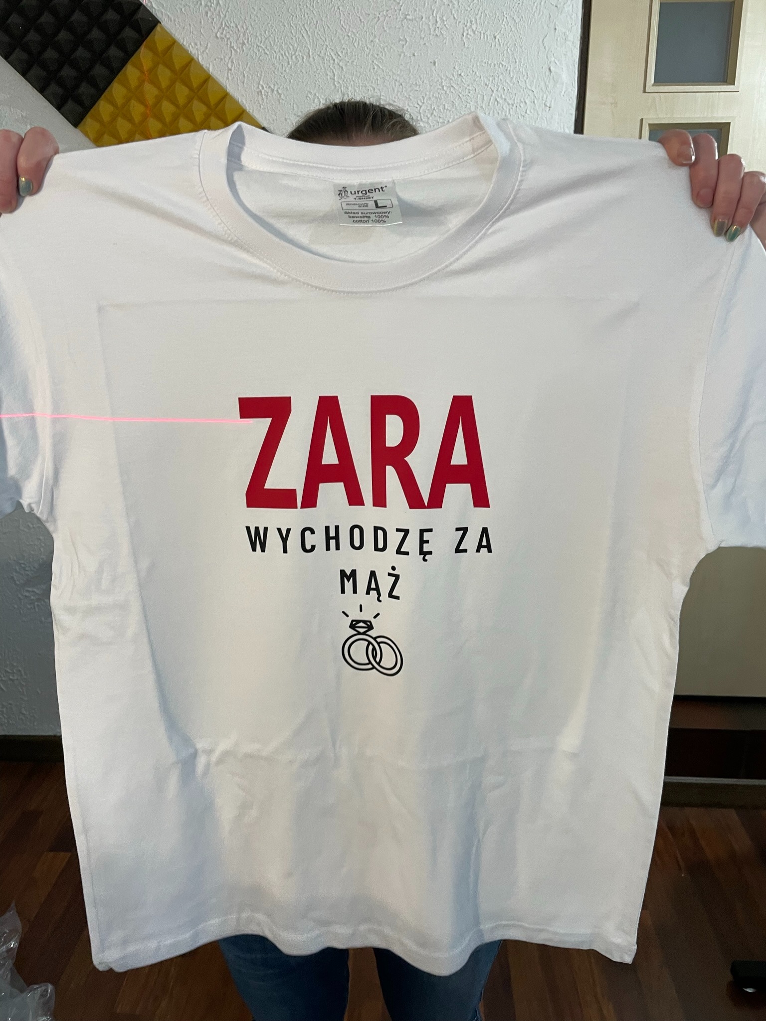 Biała koszulka z czerwonym napisem 'ZARA Wychodzę za mąż' i grafiką obrączek, prezentowana na tle ściany z panelami akustycznymi i fragmentem podłogi.