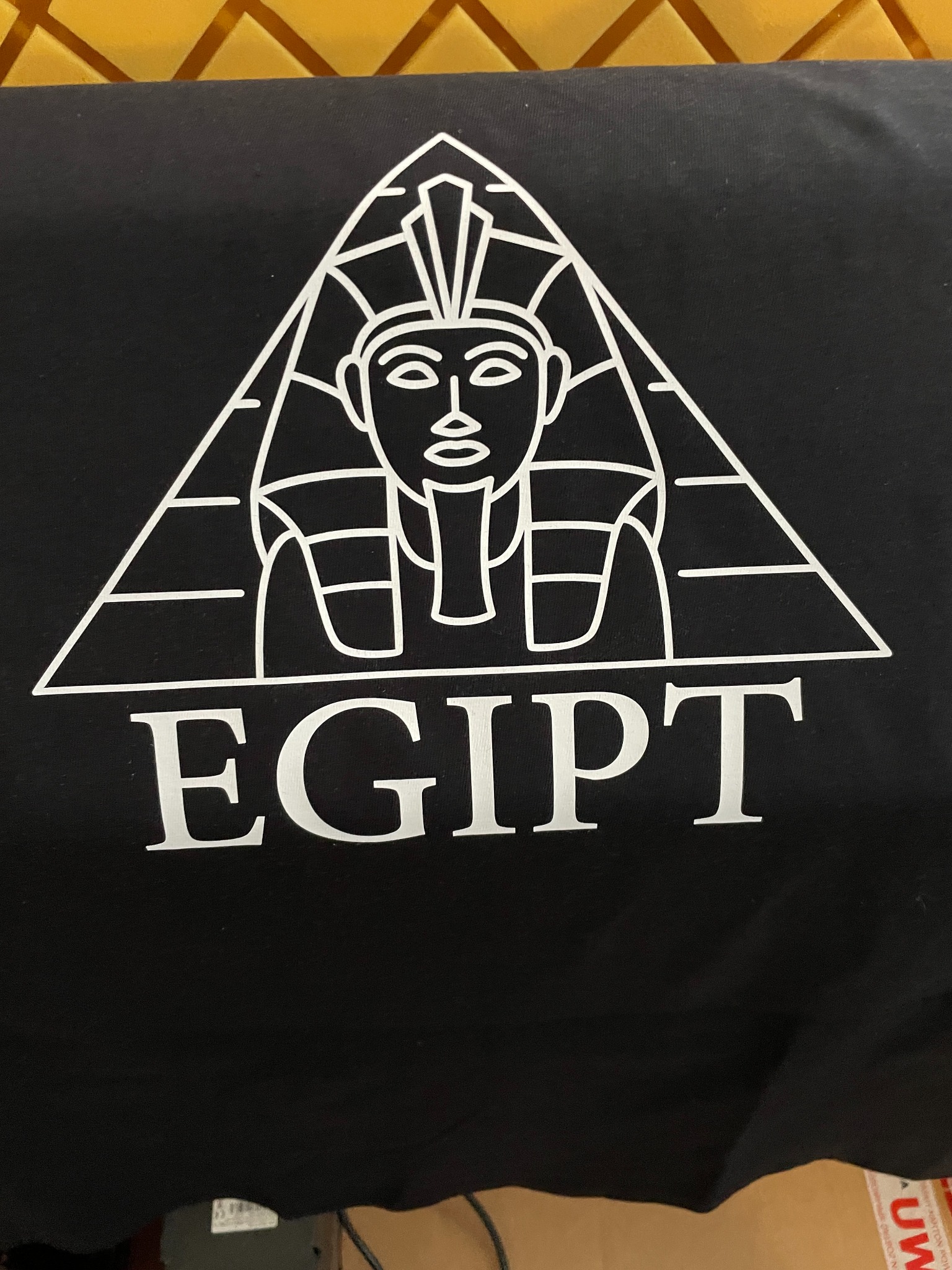Czarna koszulka z białym nadrukiem: stylizowana postać faraona wpisanego w piramidę z napisem 'EGIPT'. Nietypowa grafika na tkaninie.