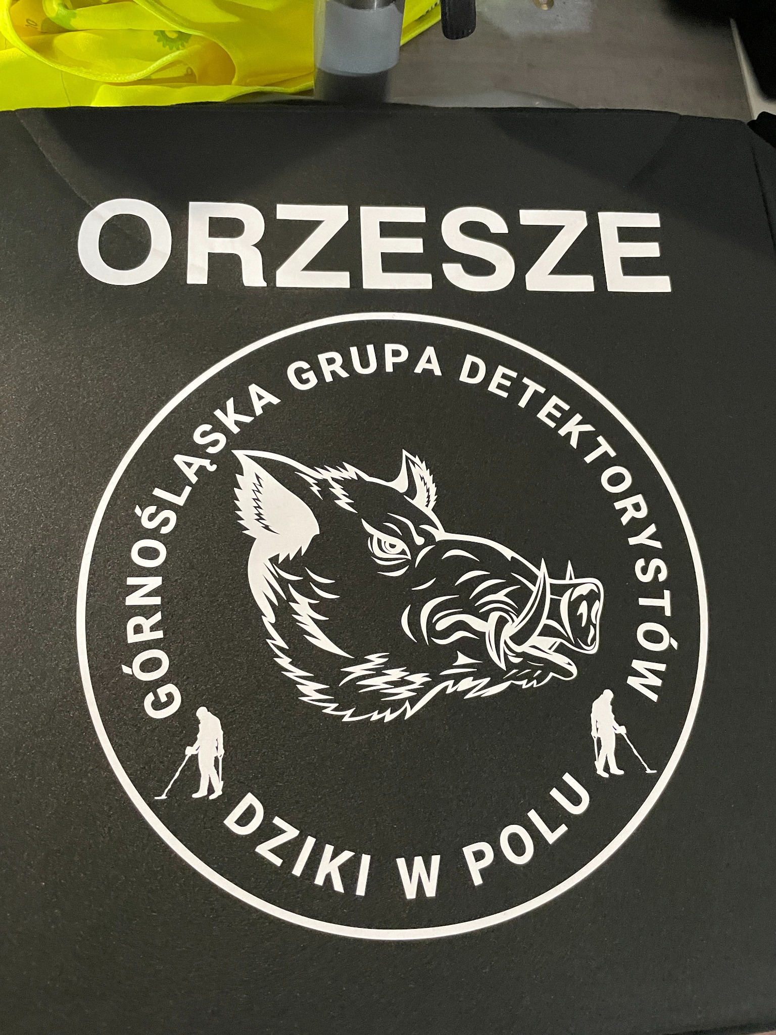 Czarna koszulka z białym nadrukiem: napis 'ORZESZE', logo 'Górnośląska Grupa Detektorystów Dzik w Polu' z wizerunkiem dzika i sylwetkami osób z wykrywaczami metali.