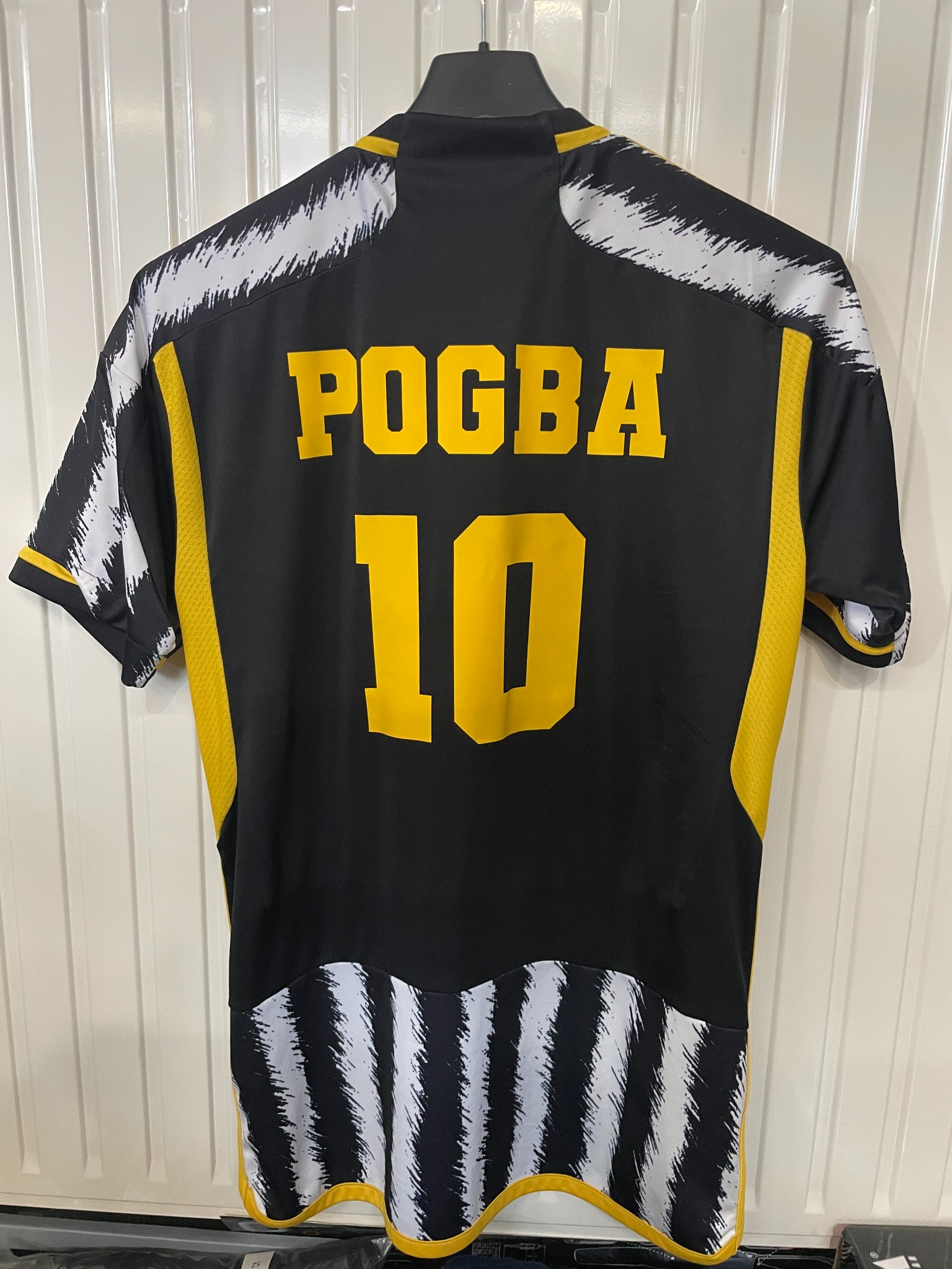 Czarno-żółta koszulka sportowa z nadrukiem 'POGBA 10' wisi na wieszaku na tle białych drzwi. Nietypowy wzór na ramionach i dole koszulki.