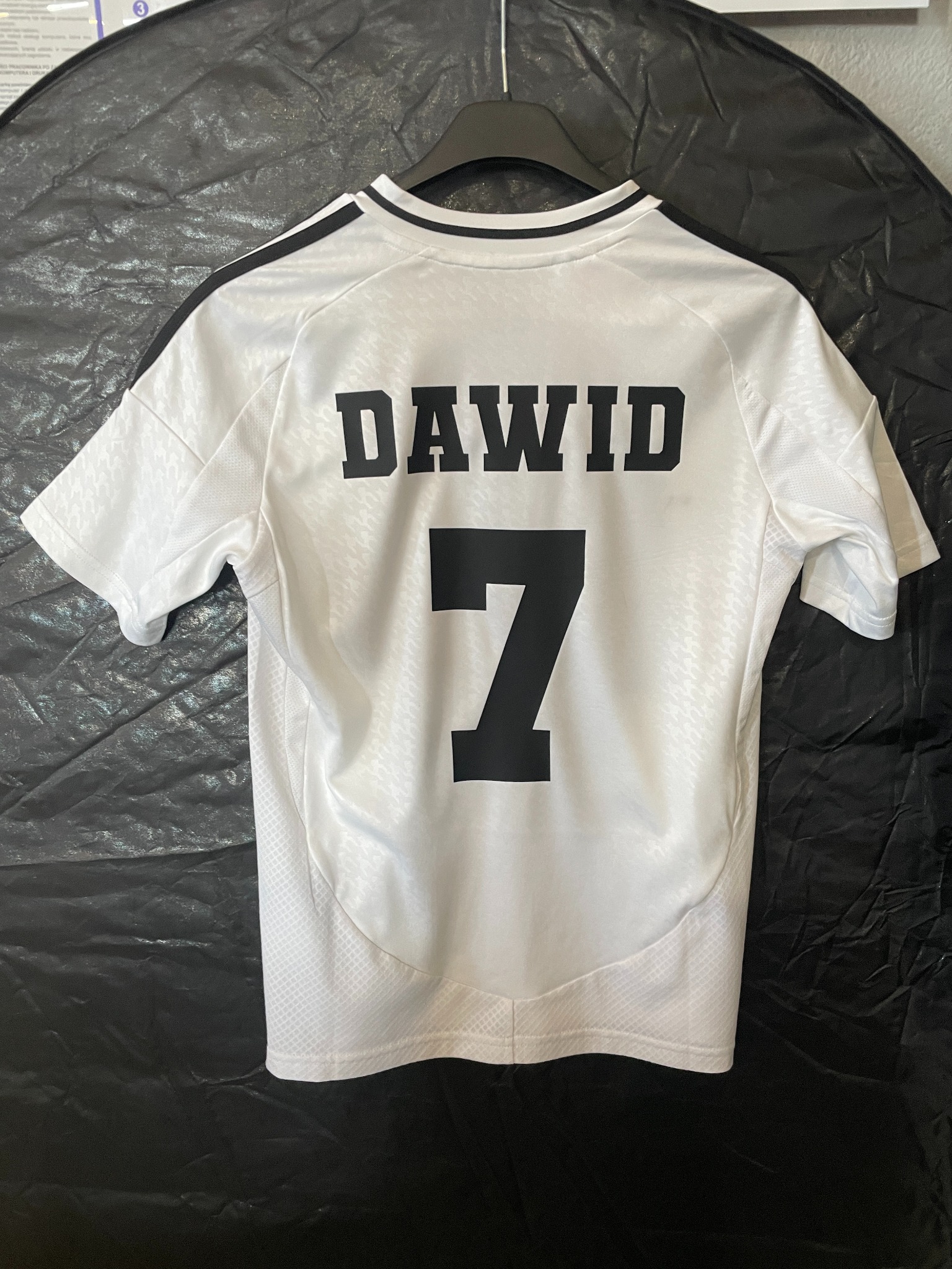 Biała koszulka sportowa z czarnym nadrukiem 'DAWID' i numerem '7', zawieszona na wieszaku. Widok z tyłu, na tle ciemnej, błyszczącej tkaniny.