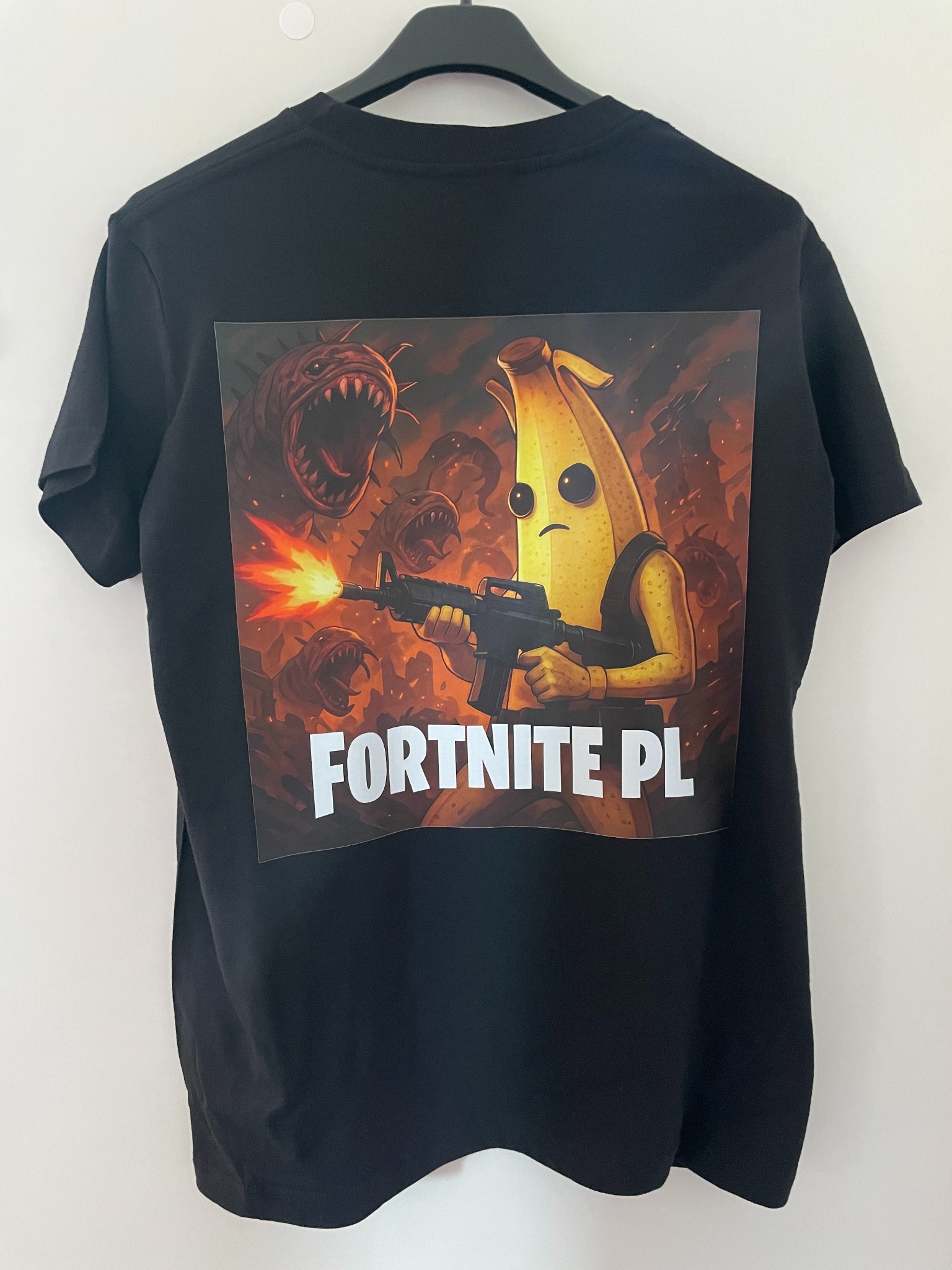 Czarna koszulka z nadrukiem postaci z gry Fortnite PL, wisząca na wieszaku na tle białej ściany. Grafika w tonacji pomarańczowo-brązowej.