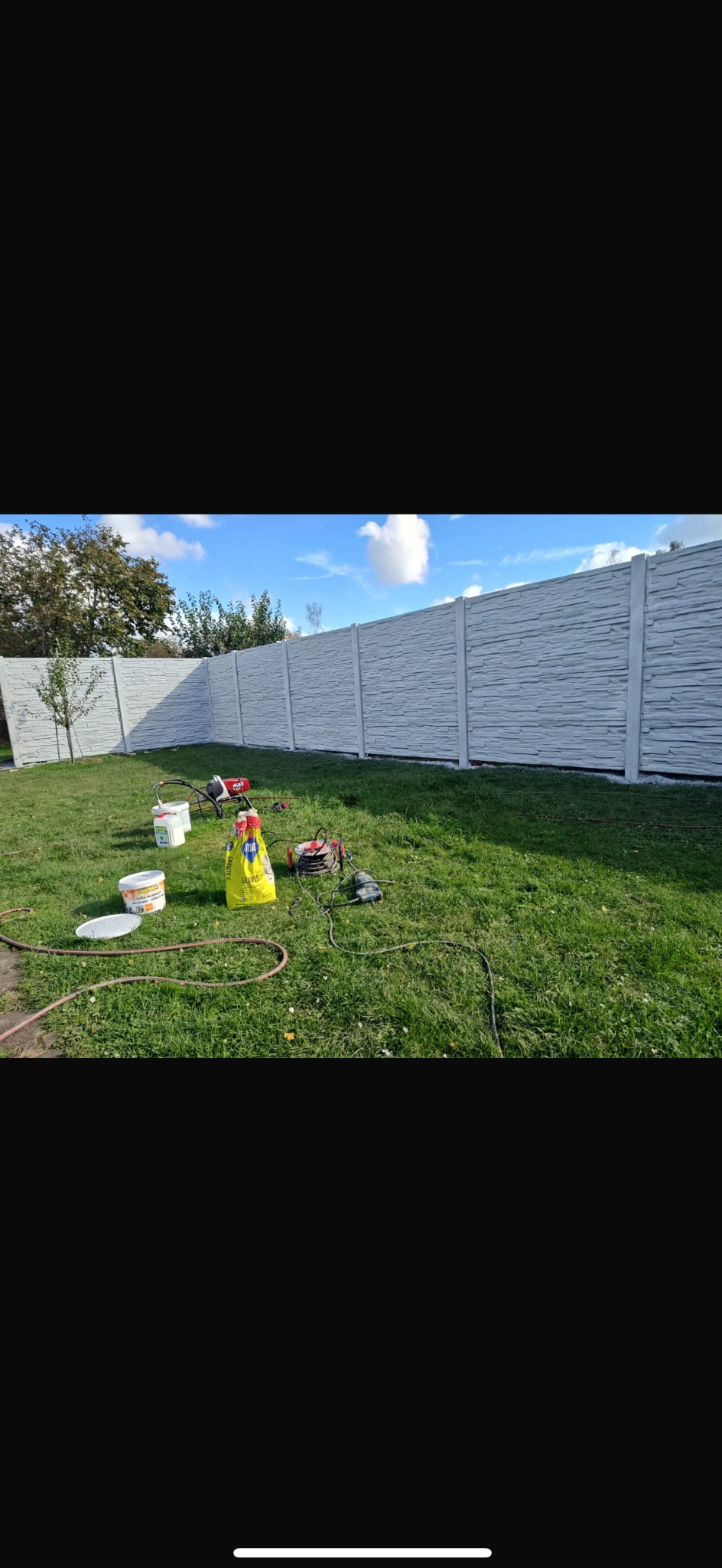 Długi, szary płot betonowy z fakturą kamienia, rozciągający się wzdłuż zielonego trawnika. Widoczne narzędzia i materiały budowlane sugerują montaż lub malowanie.