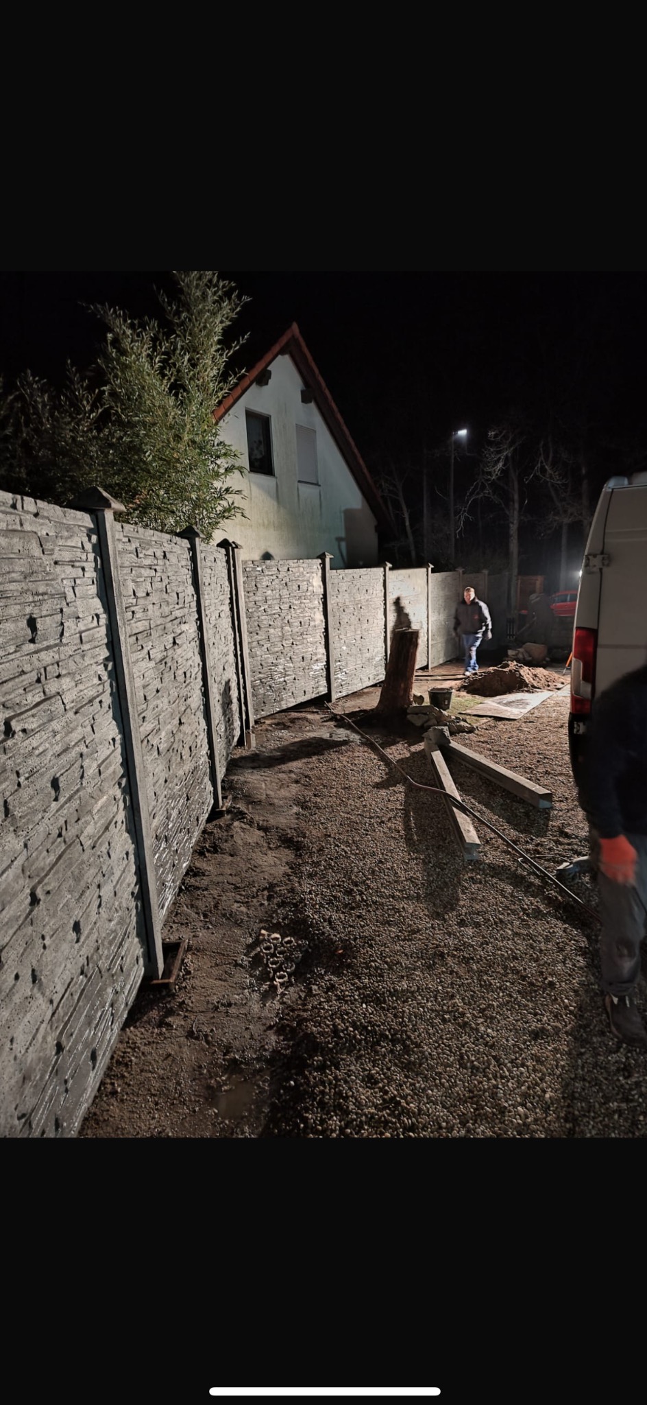 Montaż ogrodzenia betonowego imitującego kamień, widoczne panele i słupki, w tle dom jednorodzinny i pracownik w trakcie pracy.