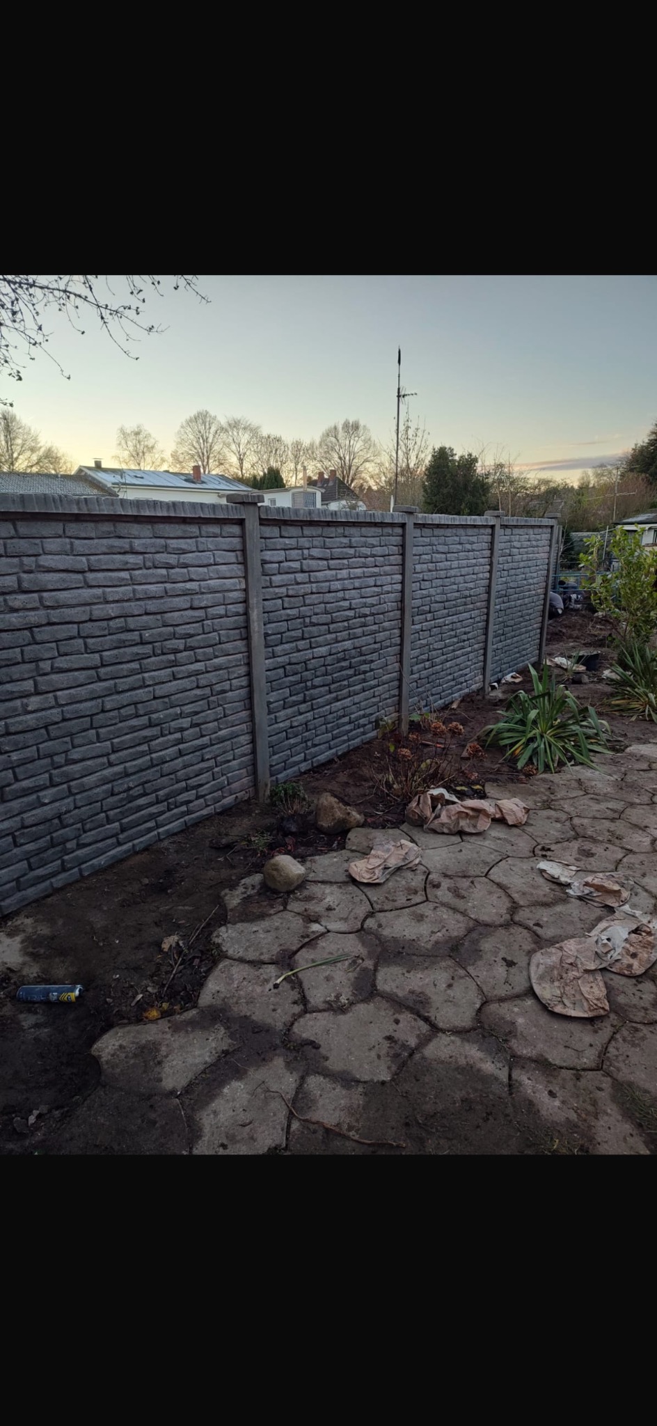 Nowoczesne, szare ogrodzenie betonowe imitujące cegłę, z widocznymi słupkami, na tle ogrodu i nieba o zmierzchu. Na pierwszym planie nieregularna, betonowa nawierzchnia.