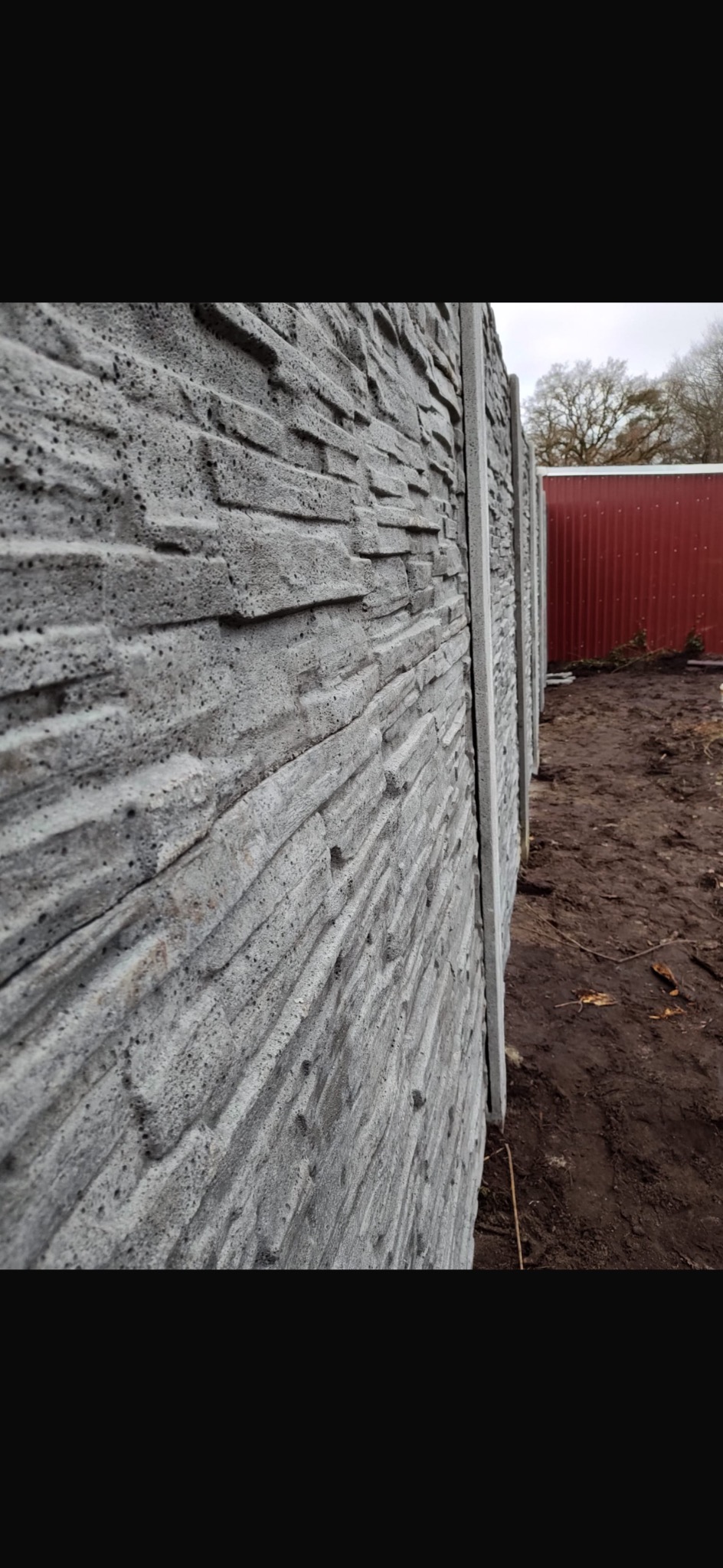 Nowoczesne ogrodzenie betonowe imitujące kamień, widok z boku, z fragmentem czerwonej blaszanej ściany w tle. Szara kolorystyka, wyraźna faktura.