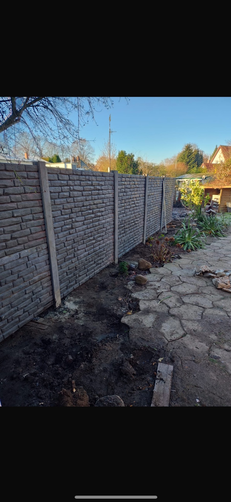 Nowoczesne ogrodzenie betonowe imitujące cegłę, z szarymi słupkami, otoczone ziemią i roślinnością, w słoneczny dzień.