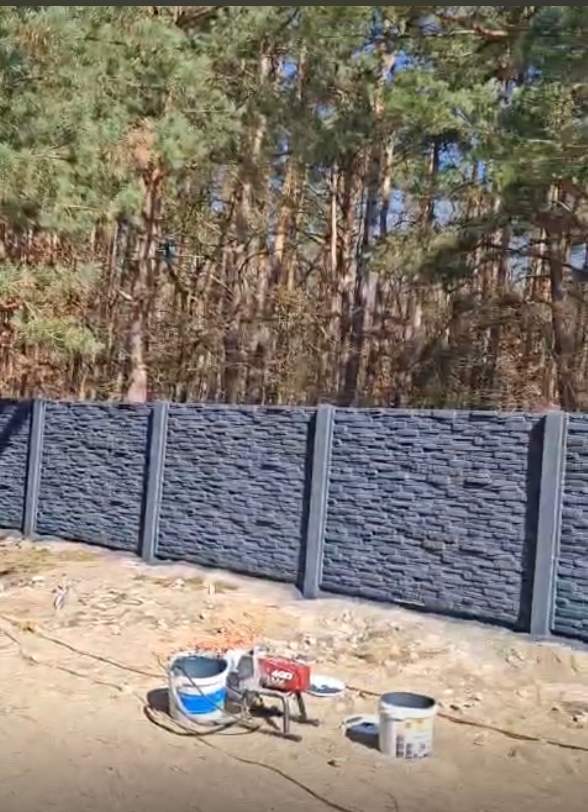 Nowoczesne ogrodzenie betonowe imitujące kamień, z szarymi słupkami, na tle lasu. Widoczne narzędzia malarskie sugerują prace wykończeniowe.