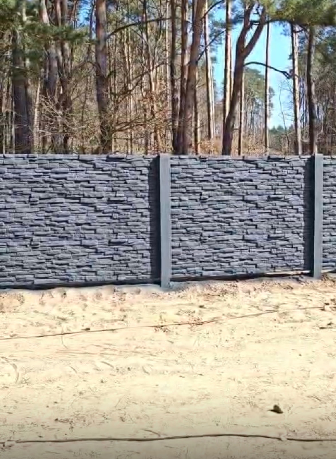 Nowoczesne ogrodzenie betonowe imitujące kamień, z szarymi słupkami, na tle lasu. Widok na gotowy fragment ogrodzenia.