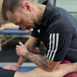 Fizjoterapeuta z tatuażem na ramieniu wykonuje masaż nogi pacjenta leżącego na stole do masażu z czerwonym wałkiem pod kolanem. Widoczne trzy paski na koszulce.