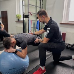 Fizjoterapia Mateusz Talarczyk - Mężczyzna w czarnej koszulce z trzema paskami pomaga klientowi wykonać ćwiczenie deski na niebieskiej piłce w siłowni. Instruktor asekuruje ćwiczącego.