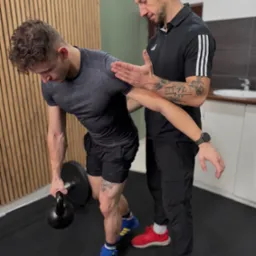 Trener personalny koryguje postawę mężczyzny podczas ćwiczenia z kettlebell. Widoczne tatuaże na ramionach obu mężczyzn, sportowe ubrania i buty w różnych kolorach.