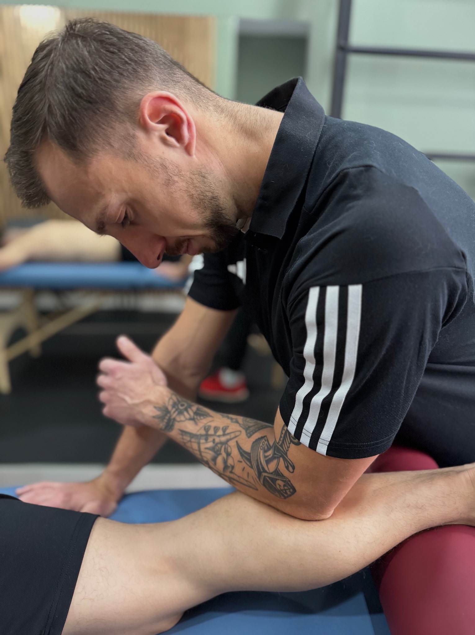 Fizjoterapeuta z tatuażem na ramieniu wykonuje masaż nogi pacjenta leżącego na stole do masażu z czerwonym wałkiem pod kolanem. Widoczne trzy paski na koszulce.
