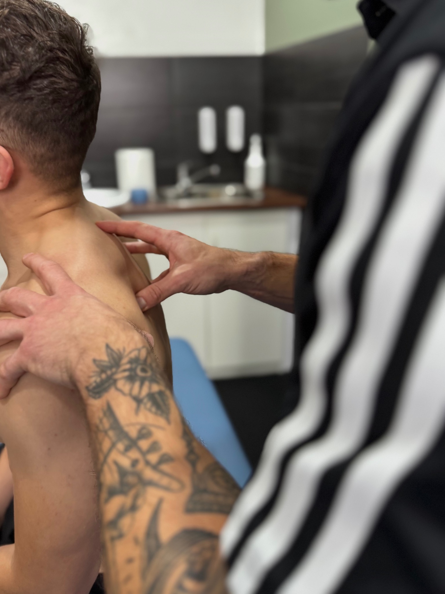Masaż ramienia pacjenta przez terapeutę w gabinecie, widoczne tatuaże na ręce terapeuty i fragment stroju w czarno-białe pasy. Tło gabinetu z umywalką.