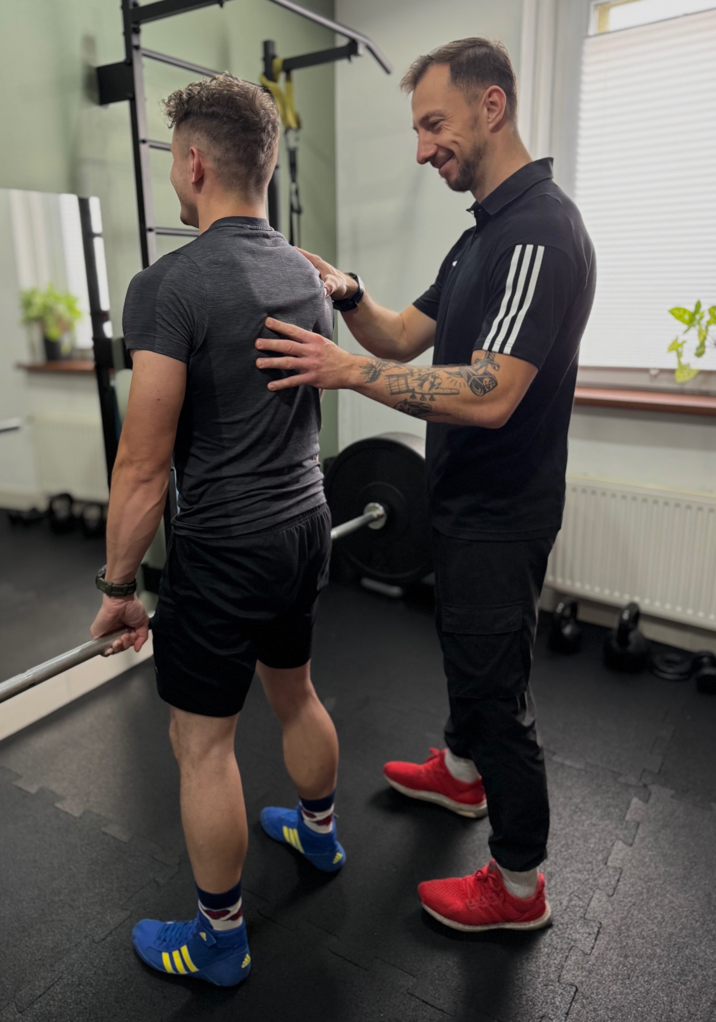 Mężczyzna z tatuażem na ramieniu koryguje postawę drugiego mężczyzny z gryfem, w sportowym stroju, na tle siłowni z lustrem i drabinką.