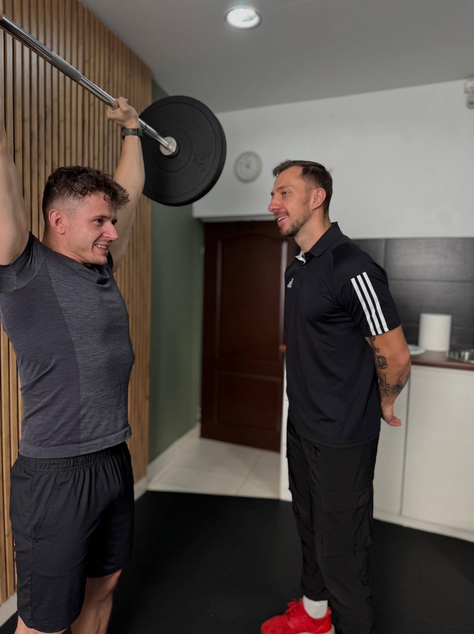 Mężczyzna ćwiczy podnoszenie sztangi pod okiem trenera w sportowym wnętrzu. Widoczny ciężar, uśmiech na twarzy ćwiczącego i skupienie trenera.