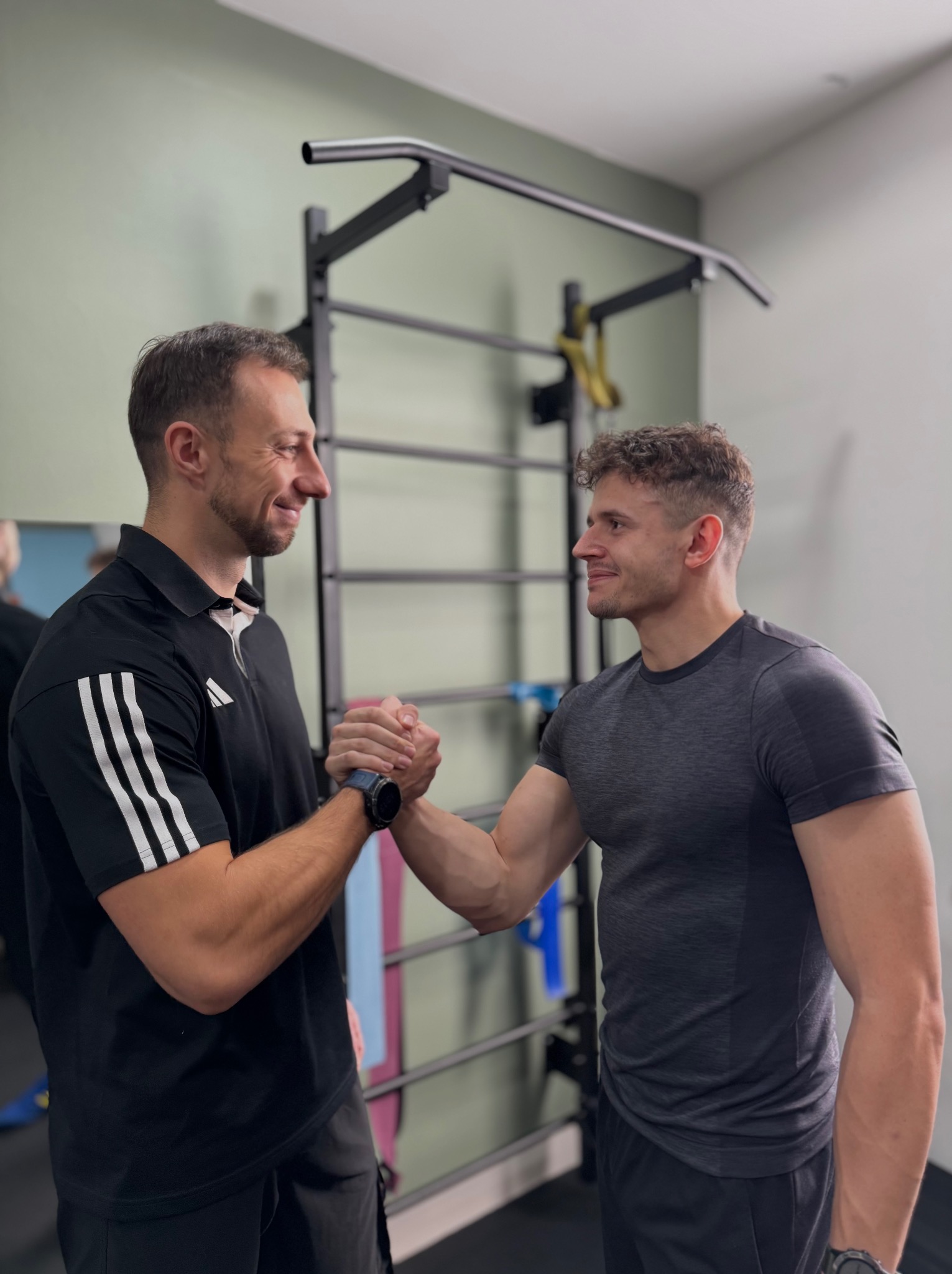 Uśmiechnięci mężczyźni ściskają sobie dłonie w siłowni, na tle drabinki gimnastycznej. Jeden w czarnej koszulce sportowej, drugi w szarej. Widoczny zegarek na ręku.