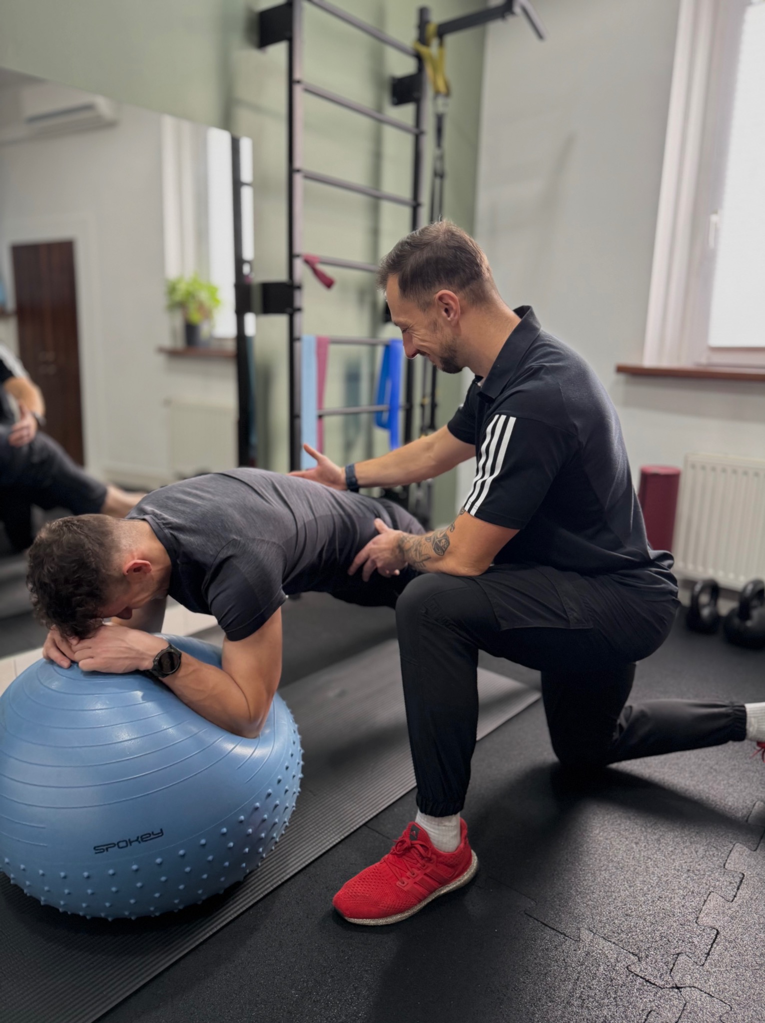 Mężczyzna w czarnej koszulce z trzema paskami pomaga klientowi wykonać ćwiczenie deski na niebieskiej piłce w siłowni. Instruktor asekuruje ćwiczącego.