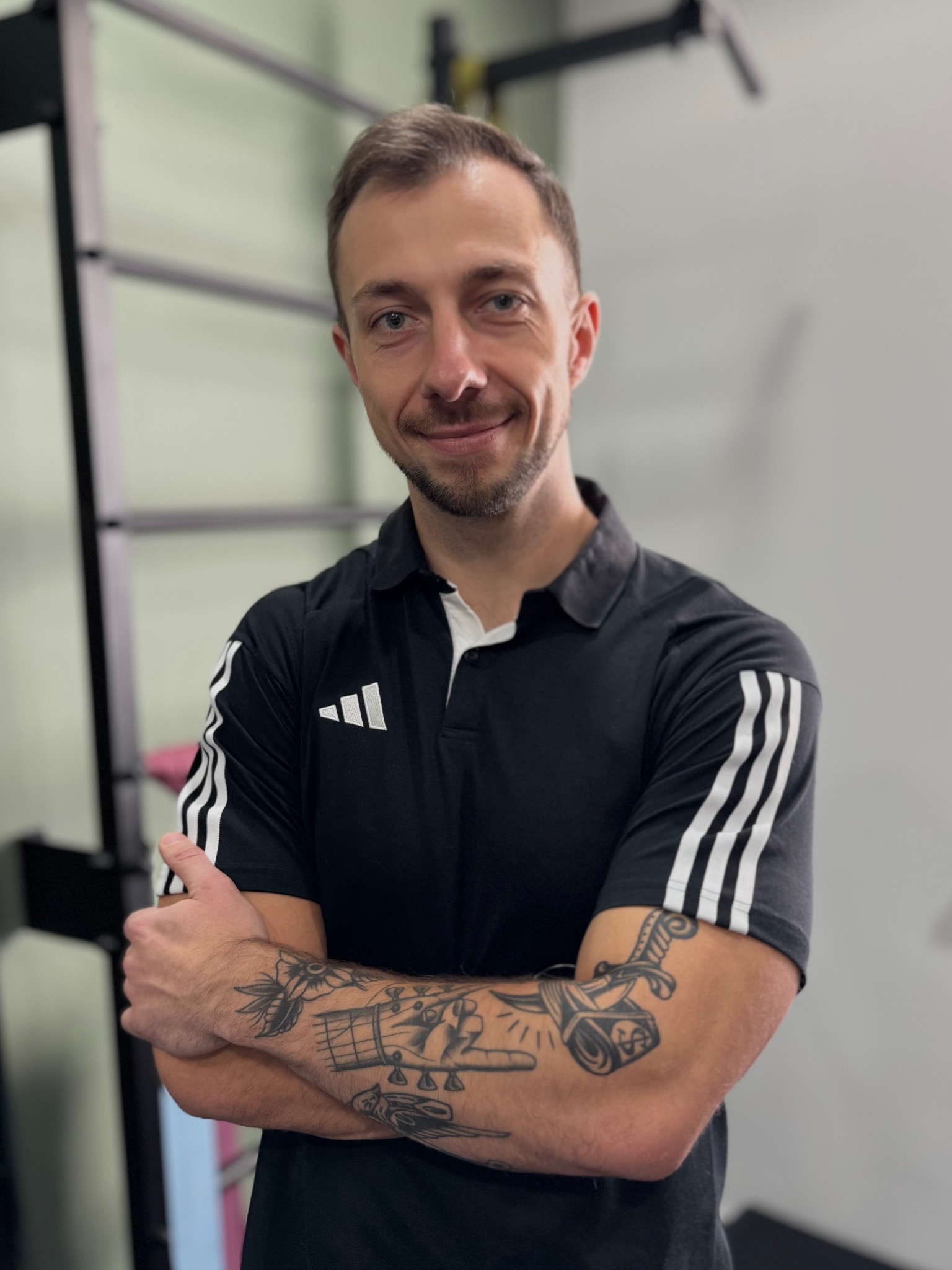 Uśmiechnięty mężczyzna w sportowej koszulce polo z tatuażami na ramionach, stoi na tle drabinki gimnastycznej w jasnym pomieszczeniu.