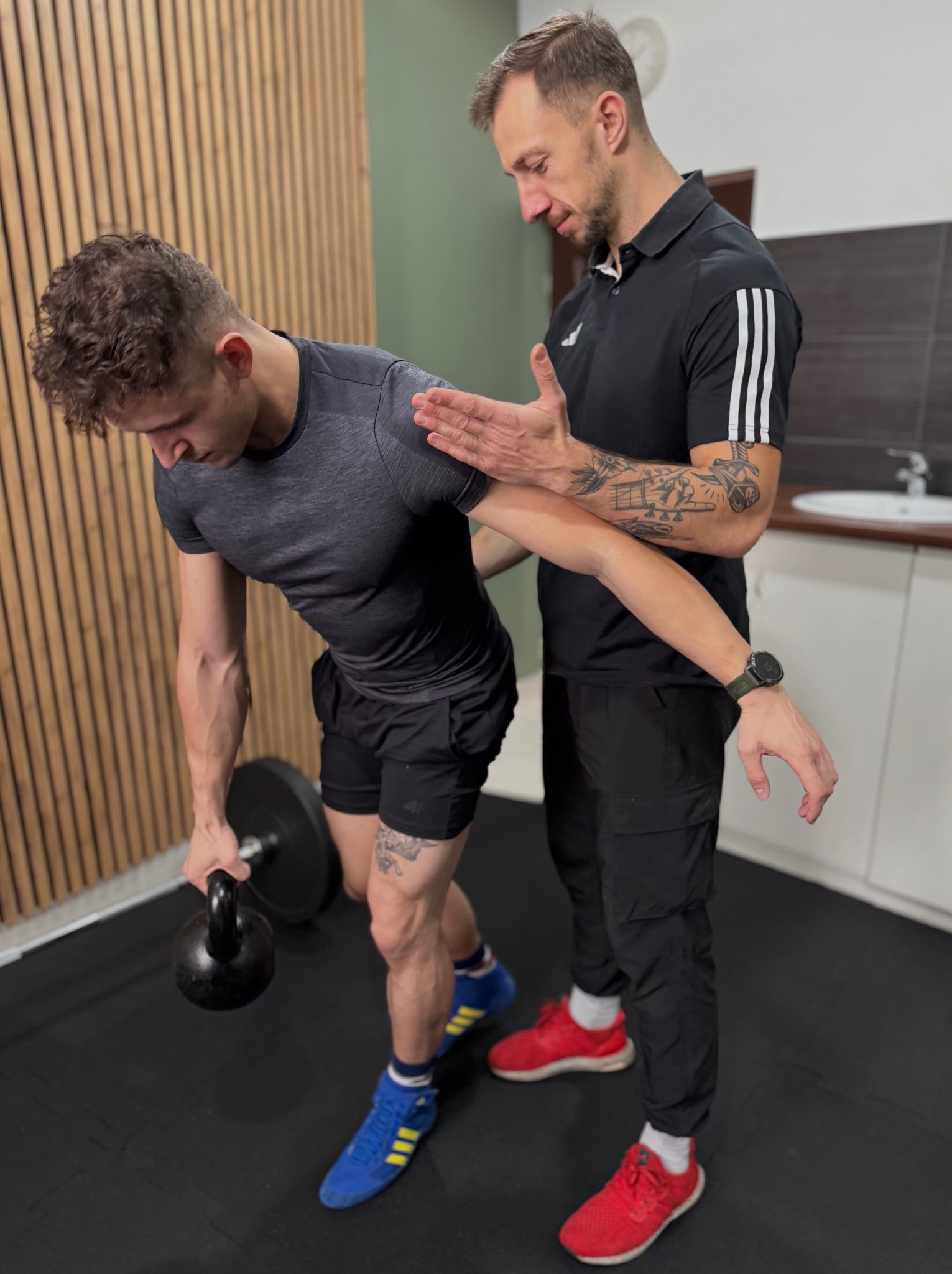 Trener personalny koryguje postawę mężczyzny podczas ćwiczenia z kettlebell. Widoczne tatuaże na ramionach obu mężczyzn, sportowe ubrania i buty w różnych kolorach.