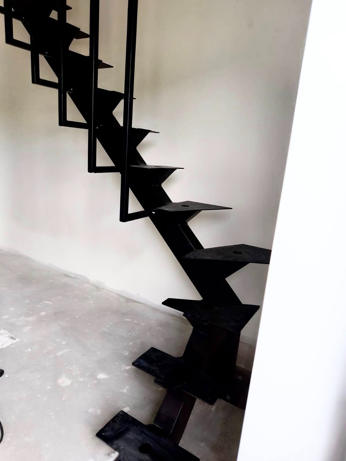 Nowoczesne, czarne schody zabiegowe z centralnym profilem i balustradą, widoczne z dołu, na tle białej ściany. Minimalistyczny design.