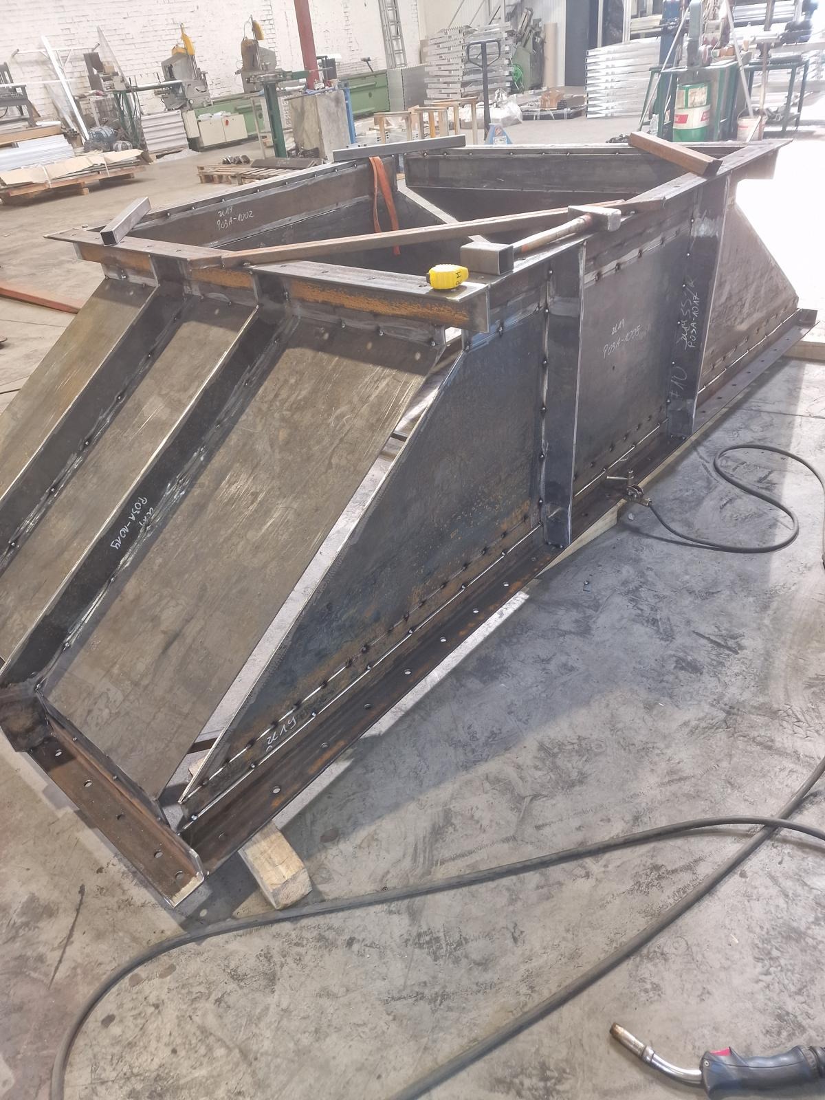 Metalowy dyfuzor w warsztacie, spawany z blachy, z widocznymi spawami i oznaczeniami. Na konstrukcji leżą narzędzia: miara i młotek. W tle warsztatowa przestrzeń.