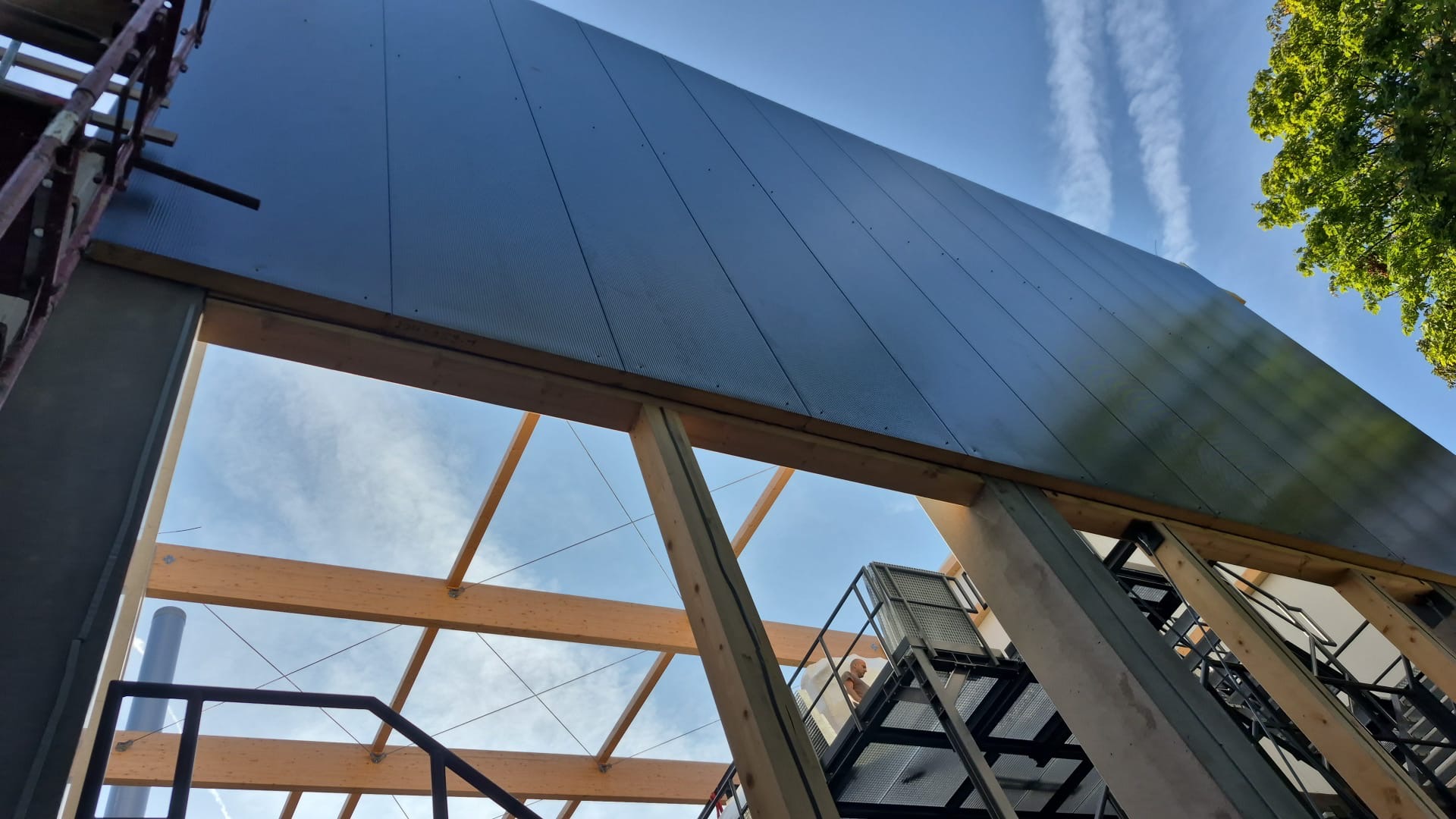 Montaż nowoczesnego, metalowego dachu z widoczną drewnianą konstrukcją nośną i rusztowaniami w słoneczny dzień. Widok z dołu na budynek w trakcie budowy.