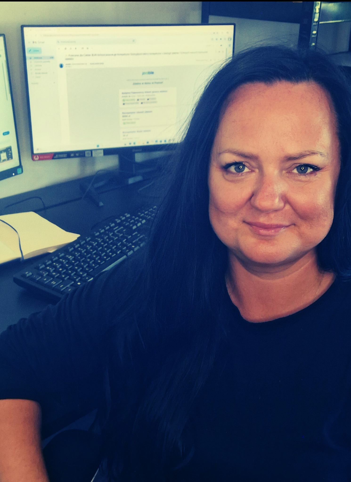 Uśmiechnięta kobieta w biurze, z komputerem i dokumentami w tle. Widok z bliska, na biurku klawiatura i segregator. Oświetlenie biurowe.