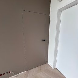 Kamil Brzazgoń Firma Budowlana ProDom - Ukryte drzwi w kolorze taupe z czarną klamką, zlicowane ze ścianą, na tle parkietu w jodełkę. Minimalistyczny design, nowoczesne wykończenie wnętrza.