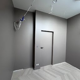 Kamil Brzazgoń Firma Budowlana ProDom - Szare wnętrze po malowaniu z parkietem w jodełkę, widoczne kable wystające ze ściany, minimalistyczne drzwi i gładkie ściany.