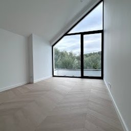 Kamil Brzazgoń Firma Budowlana ProDom - Jasne wnętrze poddasza z parkietem w jodełkę i trójkątnym oknem z widokiem na zieleń. Minimalistyczny design, świeżo po remoncie, widoczne detale wykończeniowe.