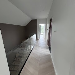 Nowoczesne wnętrze: jasny parkiet ułożony w jodełkę, szklana balustrada schodów i minimalistyczne ściany w odcieniach bieli i brązu. Widok na otwarte drzwi.