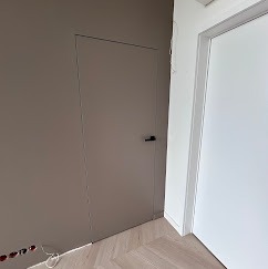 Ukryte drzwi w kolorze taupe z czarną klamką, zlicowane ze ścianą, na tle parkietu w jodełkę. Minimalistyczny design, nowoczesne wykończenie wnętrza.