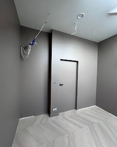 Szare wnętrze po malowaniu z parkietem w jodełkę, widoczne kable wystające ze ściany, minimalistyczne drzwi i gładkie ściany.