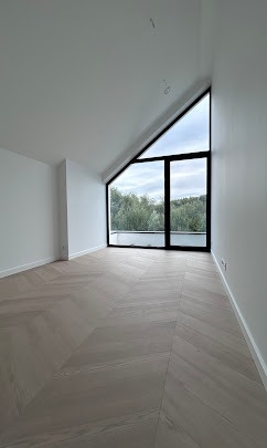 Jasne wnętrze poddasza z parkietem w jodełkę i trójkątnym oknem z widokiem na zieleń. Minimalistyczny design, świeżo po remoncie, widoczne detale wykończeniowe.